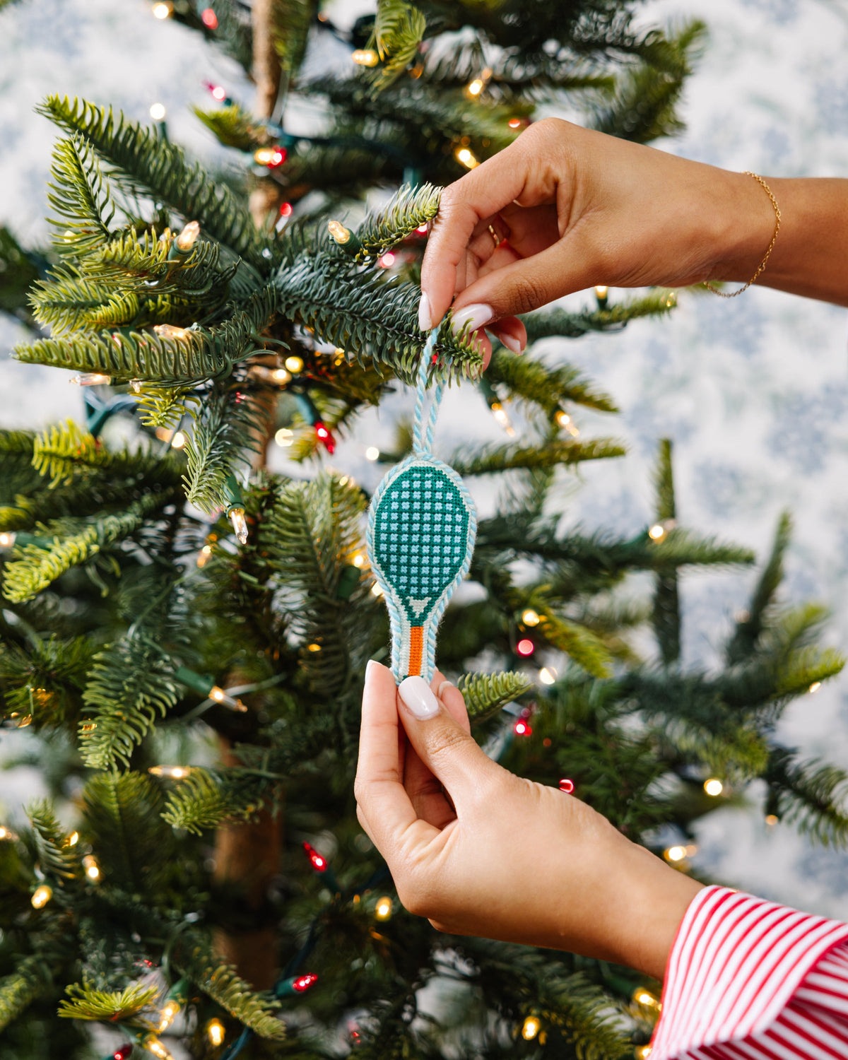 Teal needlepoint tennis racket ornament held by hand, with orange accent and loop.