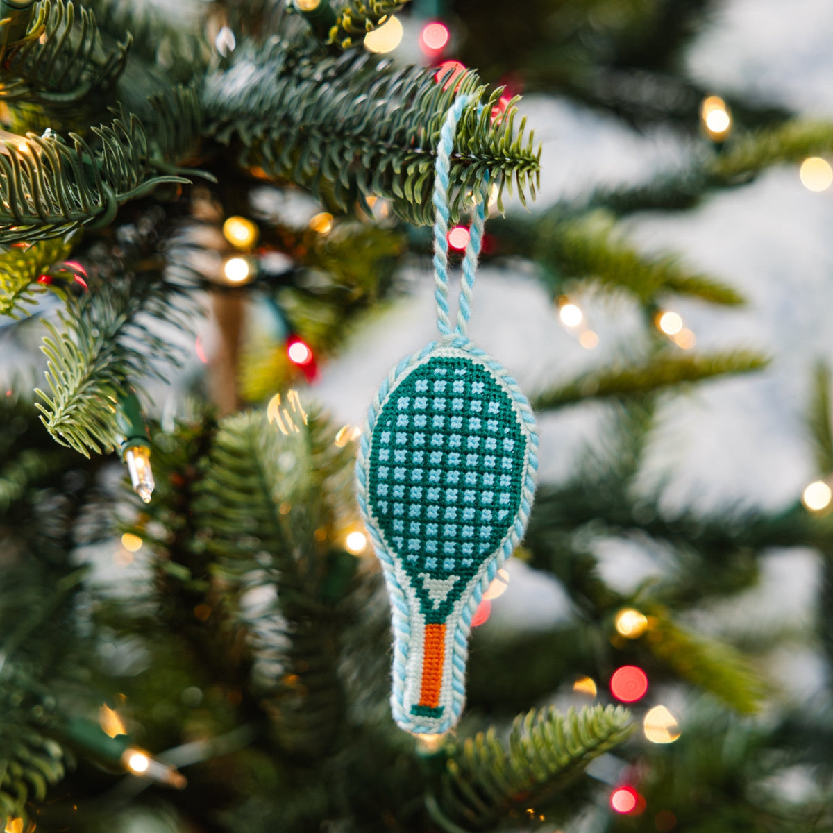 Teal needlepoint tennis racket ornament with orange handle detail and hanging loop.