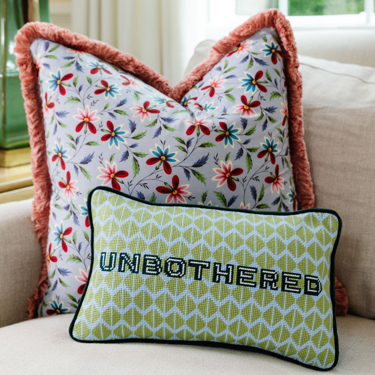 Green needlepoint throw pillow, black trim, UNBOTHERED lettering, on floral bedding
