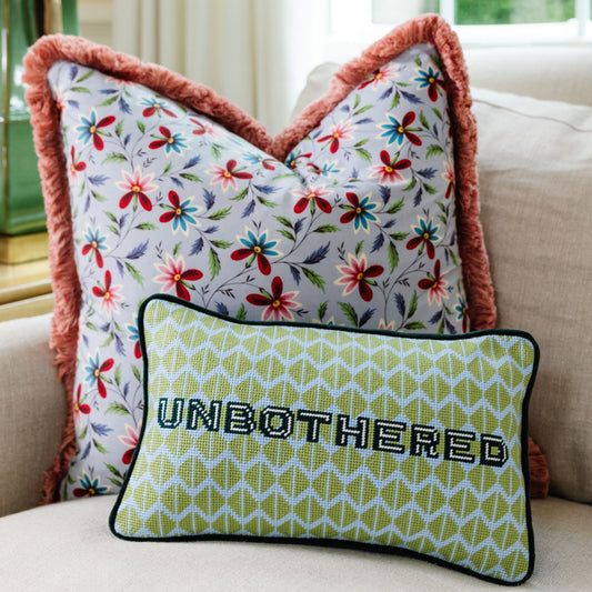 Green needlepoint throw pillow, black trim, UNBOTHERED lettering, on floral bedding