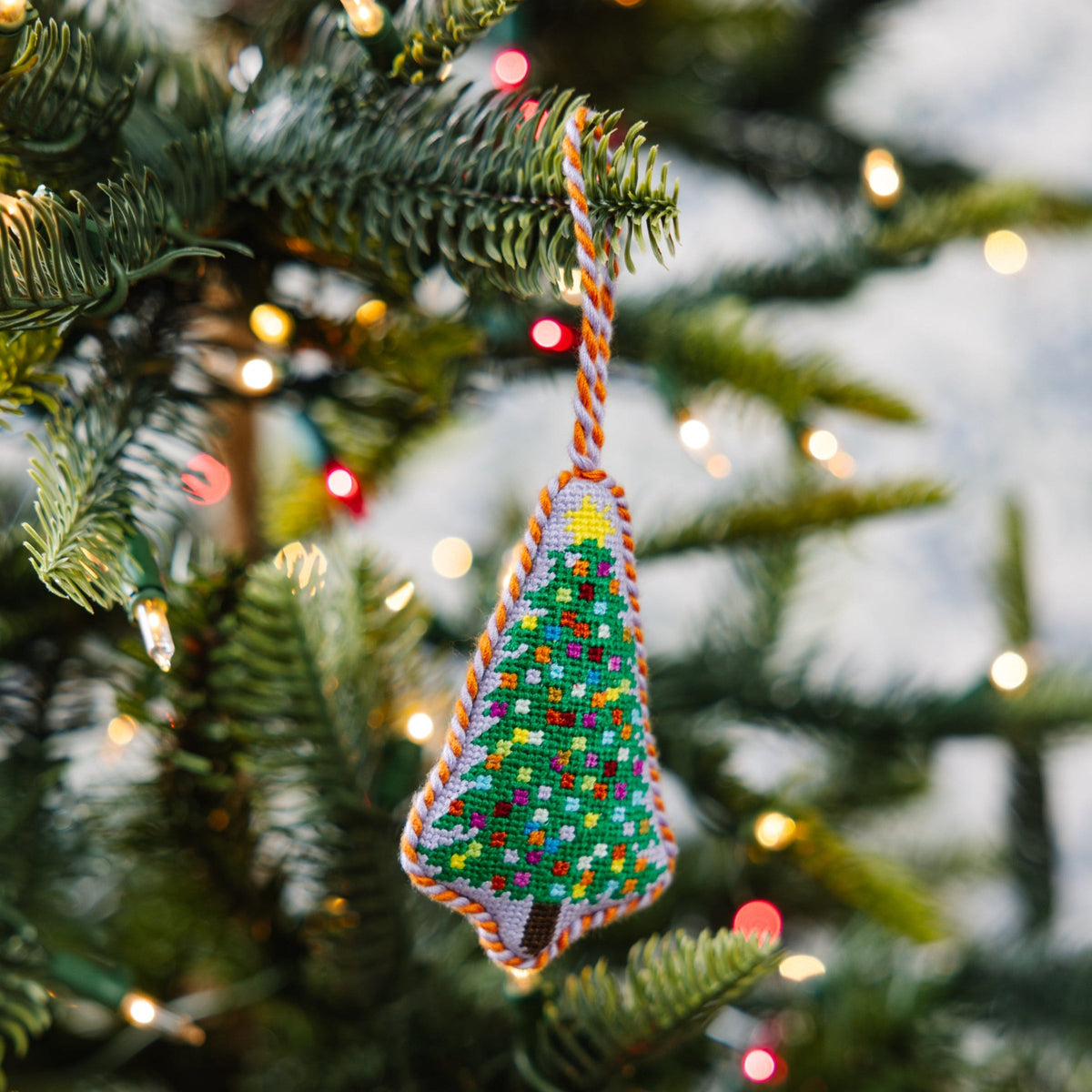 Needlepoint tree ornament hanging on evergreen branch, multicolor stitched pattern