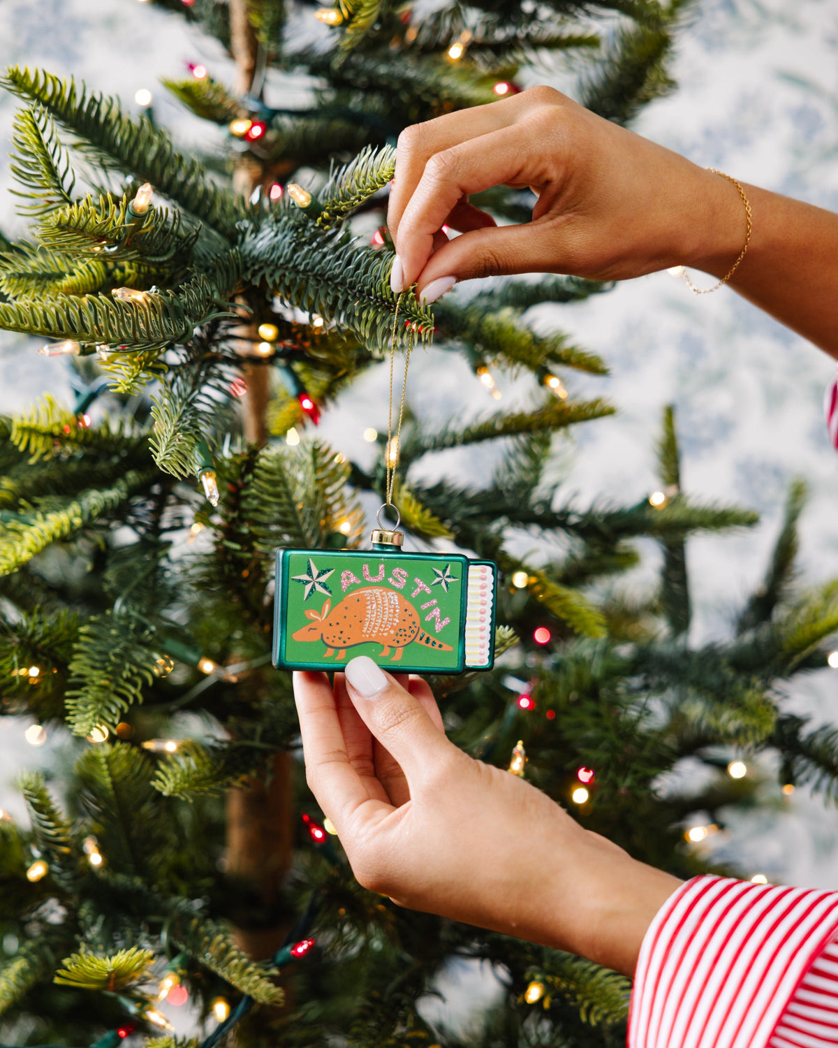 Hands placing a green Austin matchbook ornament on pine needles with gold cord