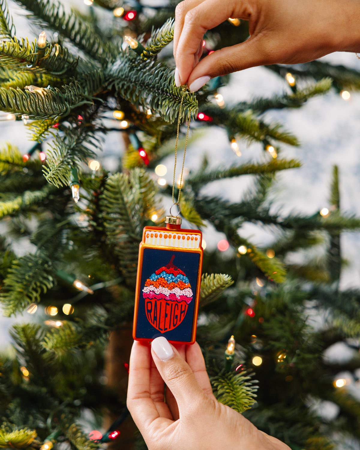 Hand holding matchbook-shaped Raleigh ornament on gold cord, orange frame details