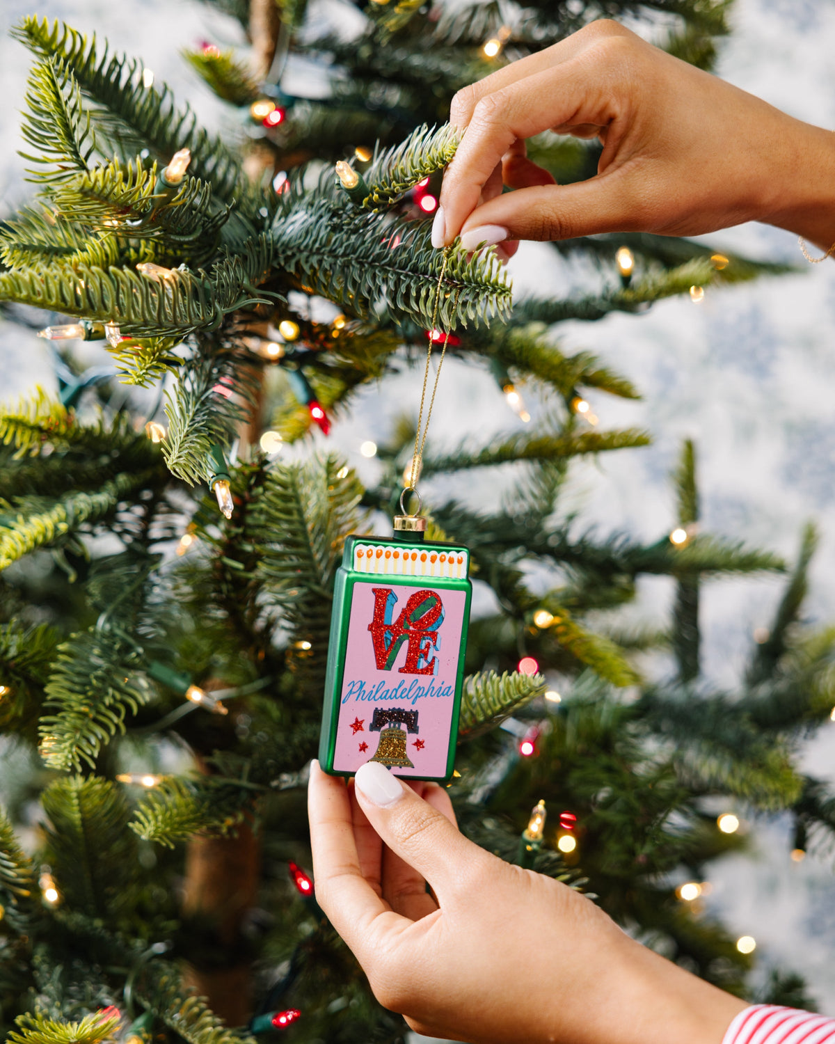 Hands placing a matchbook ornament with 'VE' and Philadelphia artwork on a tree