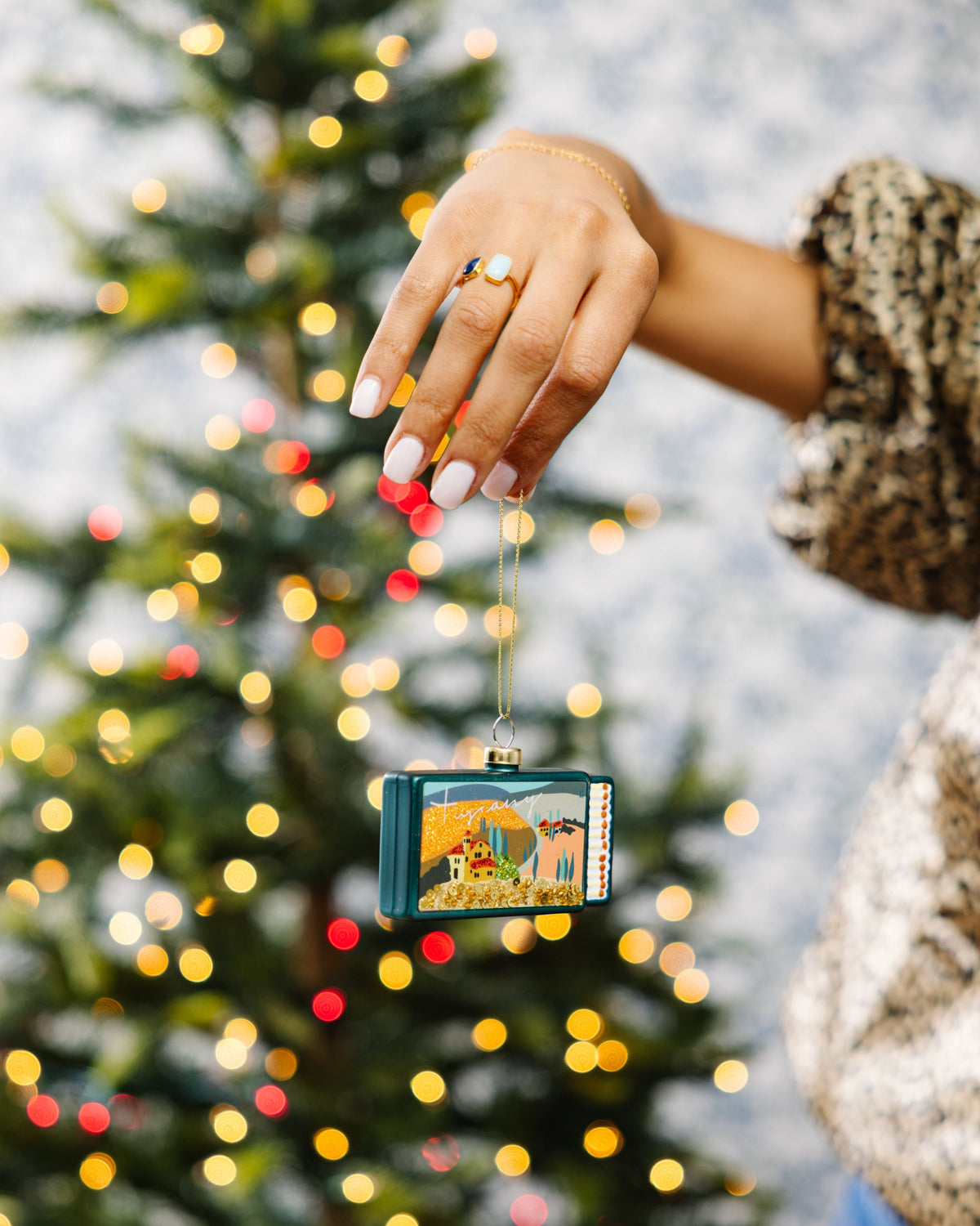 Tuscany matchbook ornament held by a hand, teal glitter landscape with gold bead trim