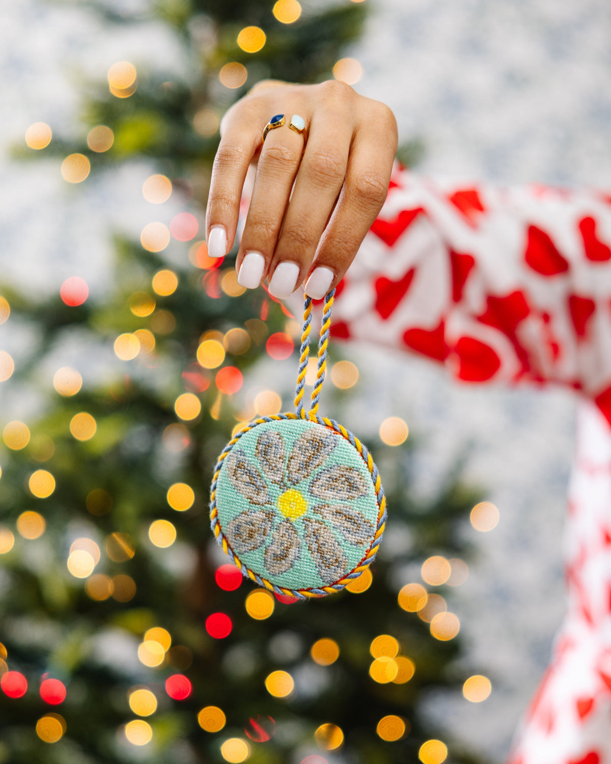Oyster needlepoint icon ornament held by a hand, teal circle with yellow center and braided cord