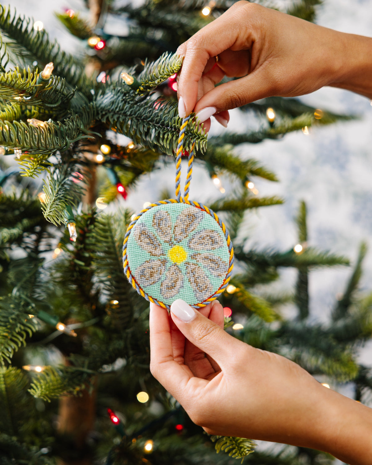 Oyster needlepoint icon ornament held by hands near tree, teal circle with yellow center