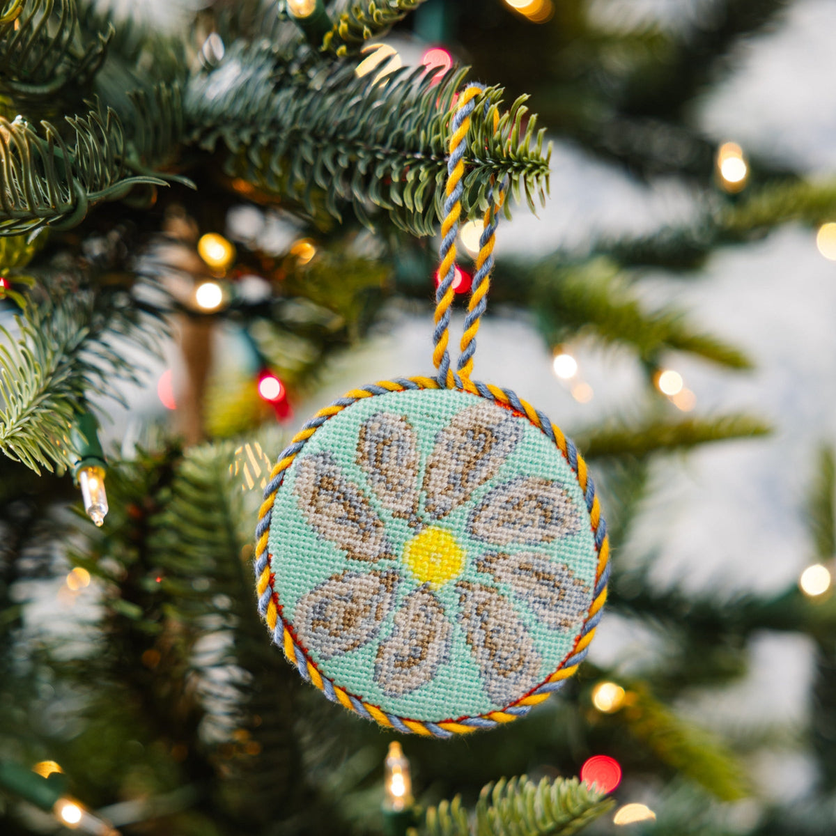 Oyster needlepoint icon ornament hanging on a tree, teal circle with yellow center and braid cord
