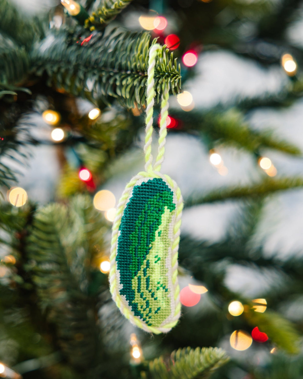 Green needlepoint pickle ornament with white border and yellow stitching, hanging loop