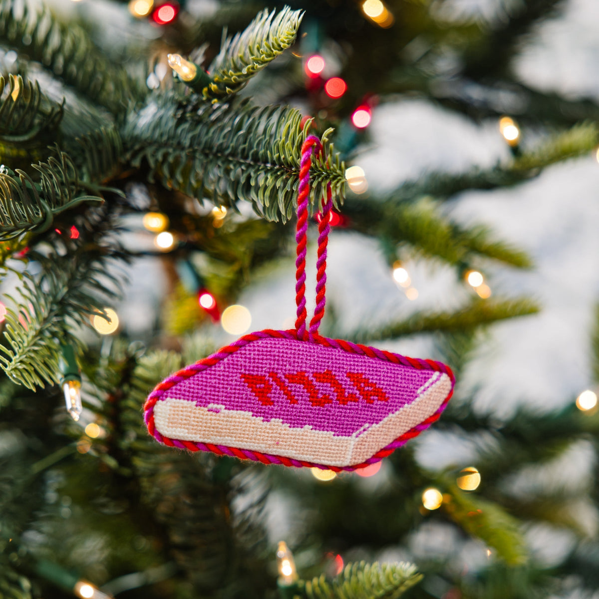 Needlepoint pizza slice ornament in magenta stitch pattern, hanging on pine branch