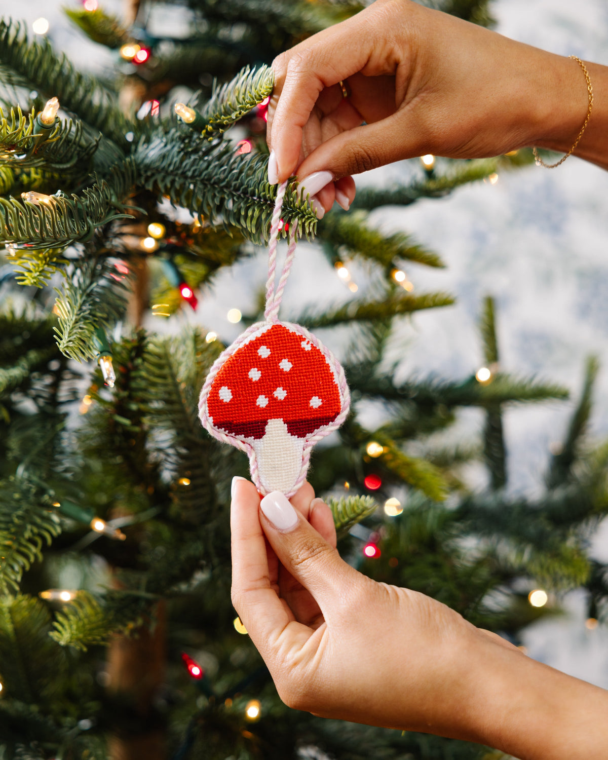 Needlepoint mushroom ornament being hung on a tree, red cap with white spots and stem