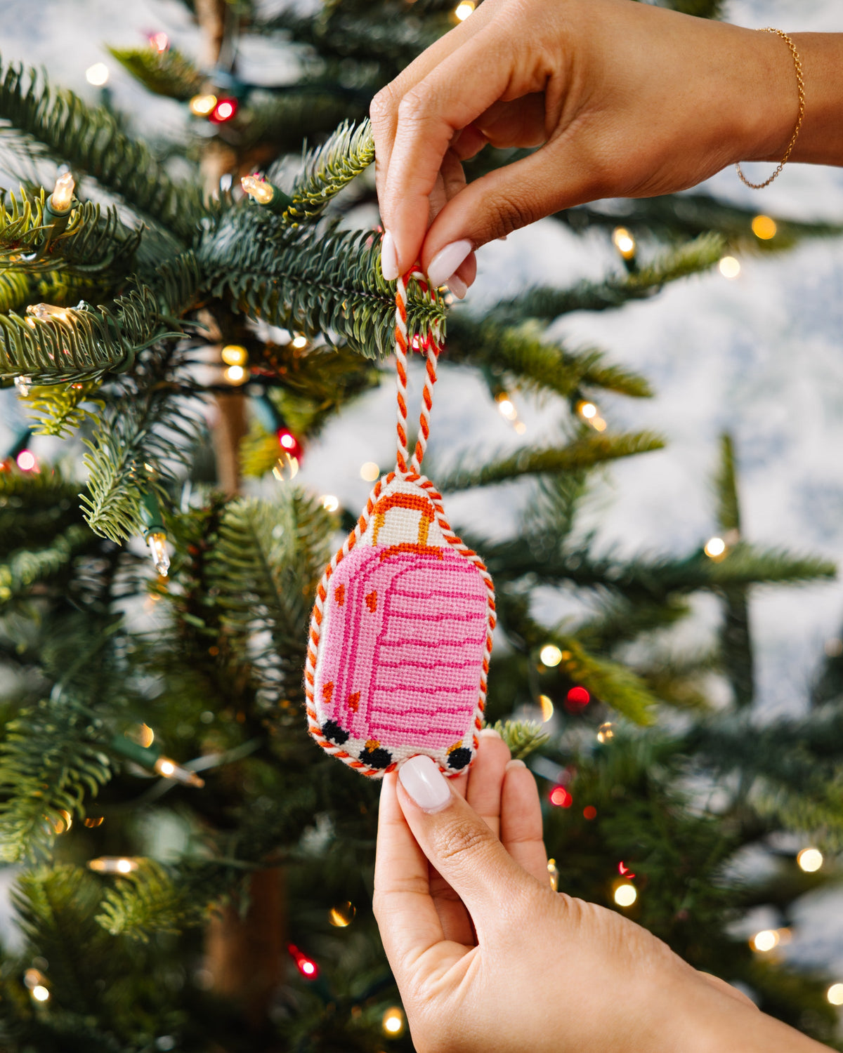 Hands attaching pink luggage needlepoint ornament to a Christmas tree branch via cord