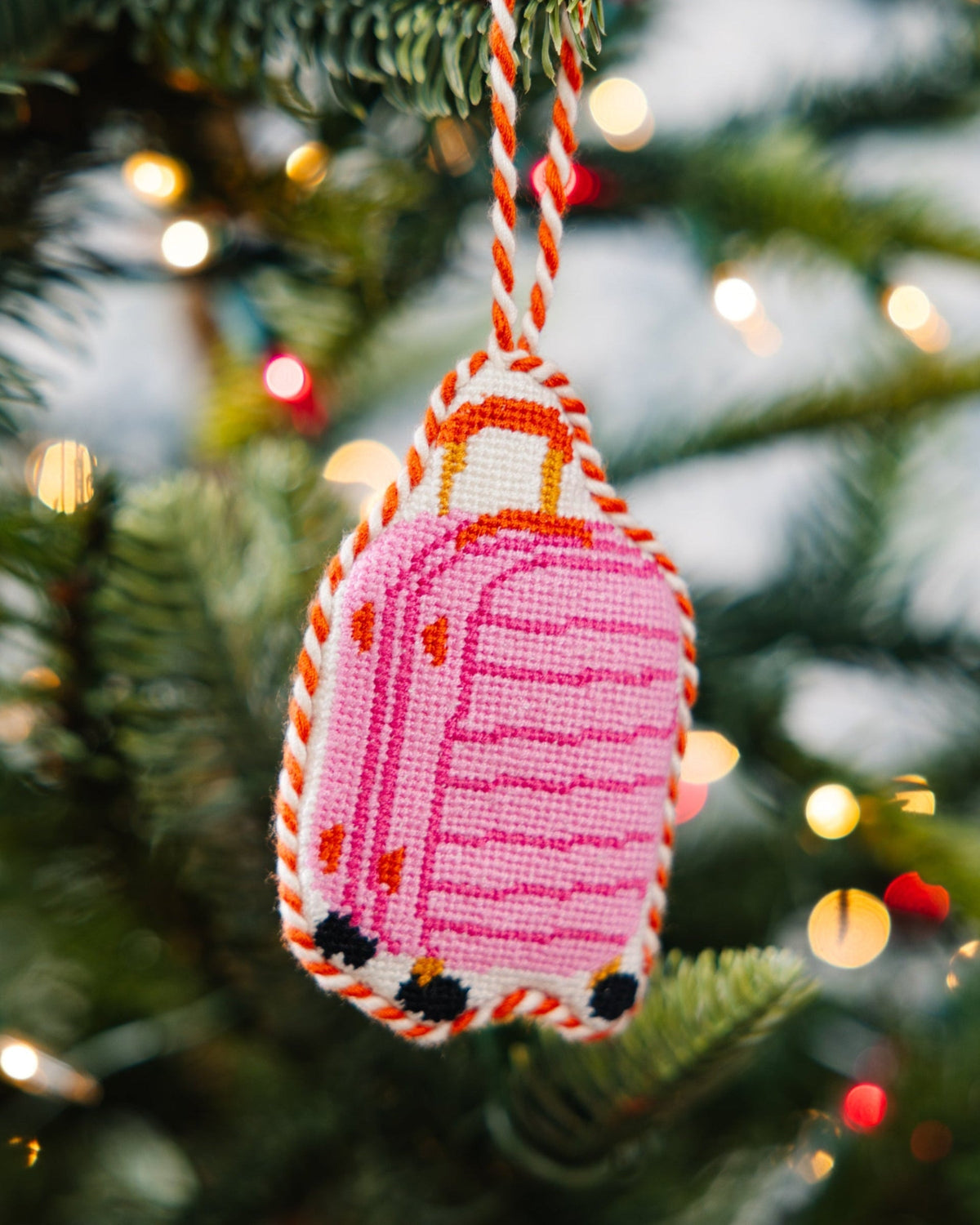 Pink luggage needlepoint ornament hanging on a white-and-orange twisted cord