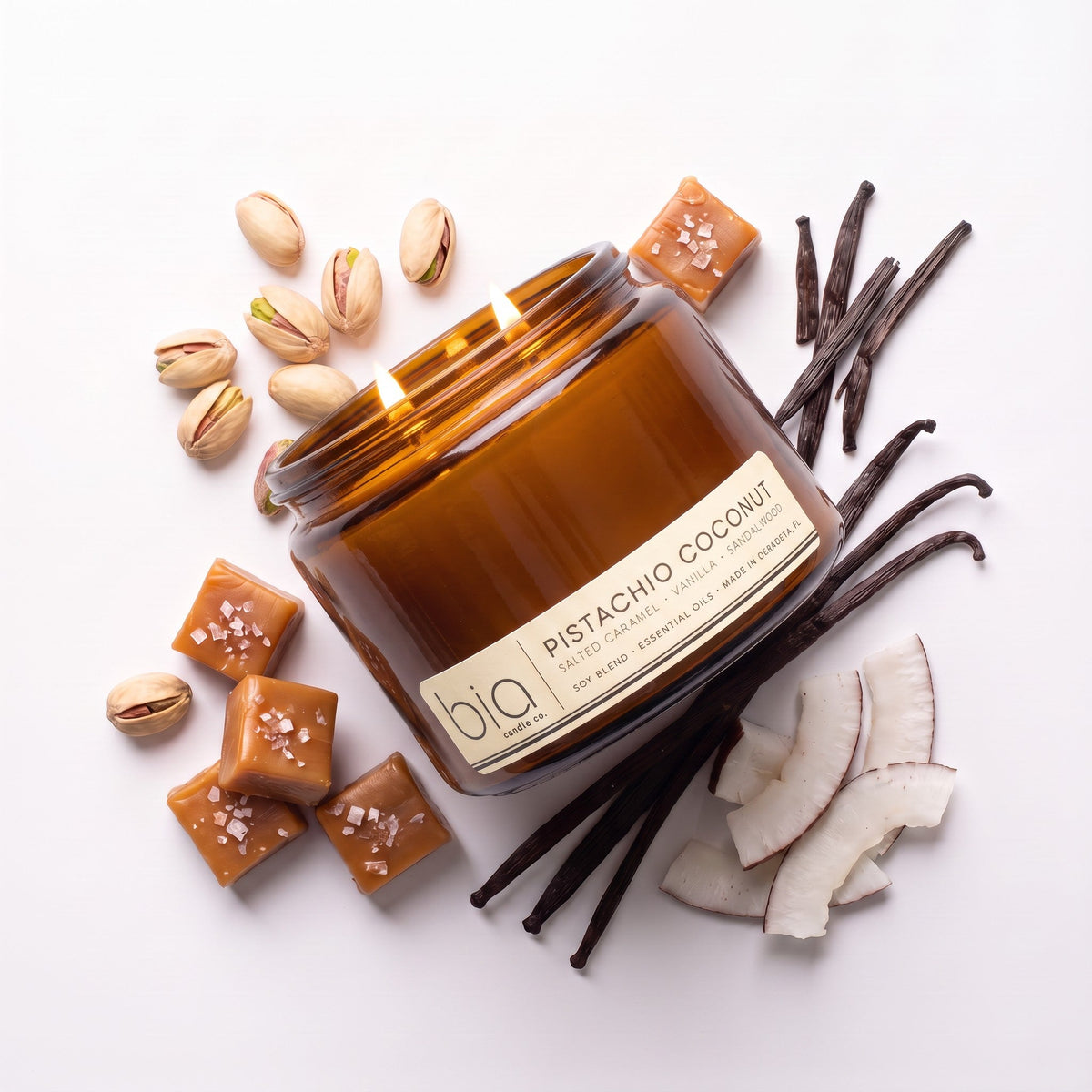 Rectangular candle jar with lit wick, surrounded by vanilla pods, pistachios, and coconut pieces.
