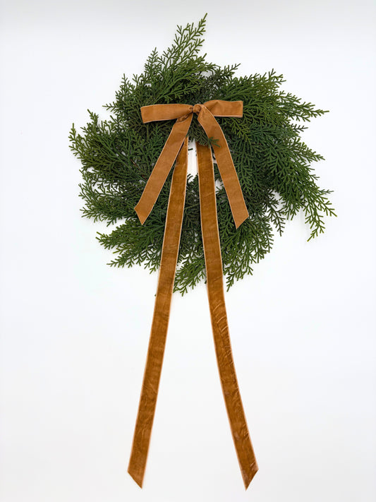 Mini cedar wreath with long tan ribbon bows hanging on a wall hook