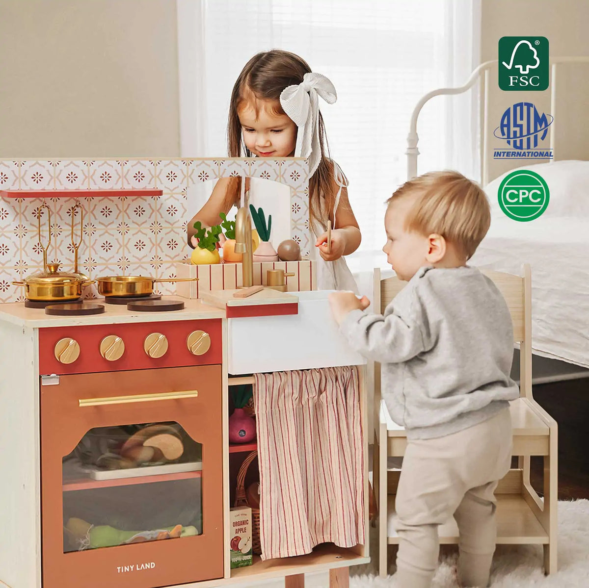 Tiny Land play kitchen with oven, stovetop, sink, and curtained storage beside two children