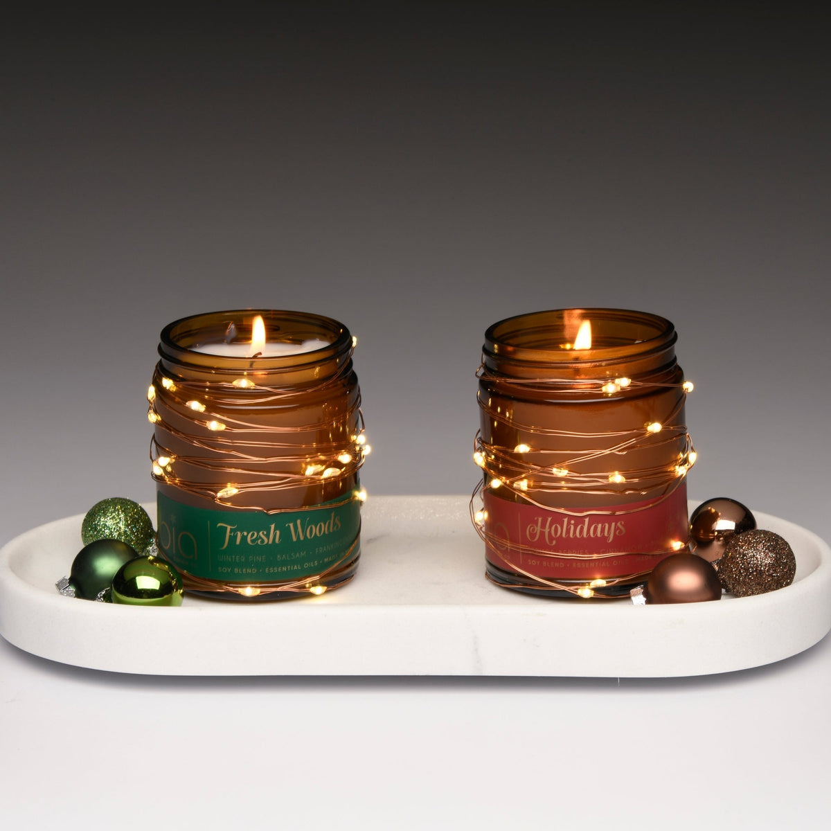Two amber glass soy candles on a white tray with green Fresh Woods and red Holidays labels, both lit