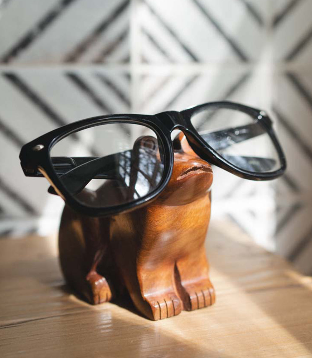 Hand-carved wooden frog eyeglass holder stand holding black eyeglasses, angled view