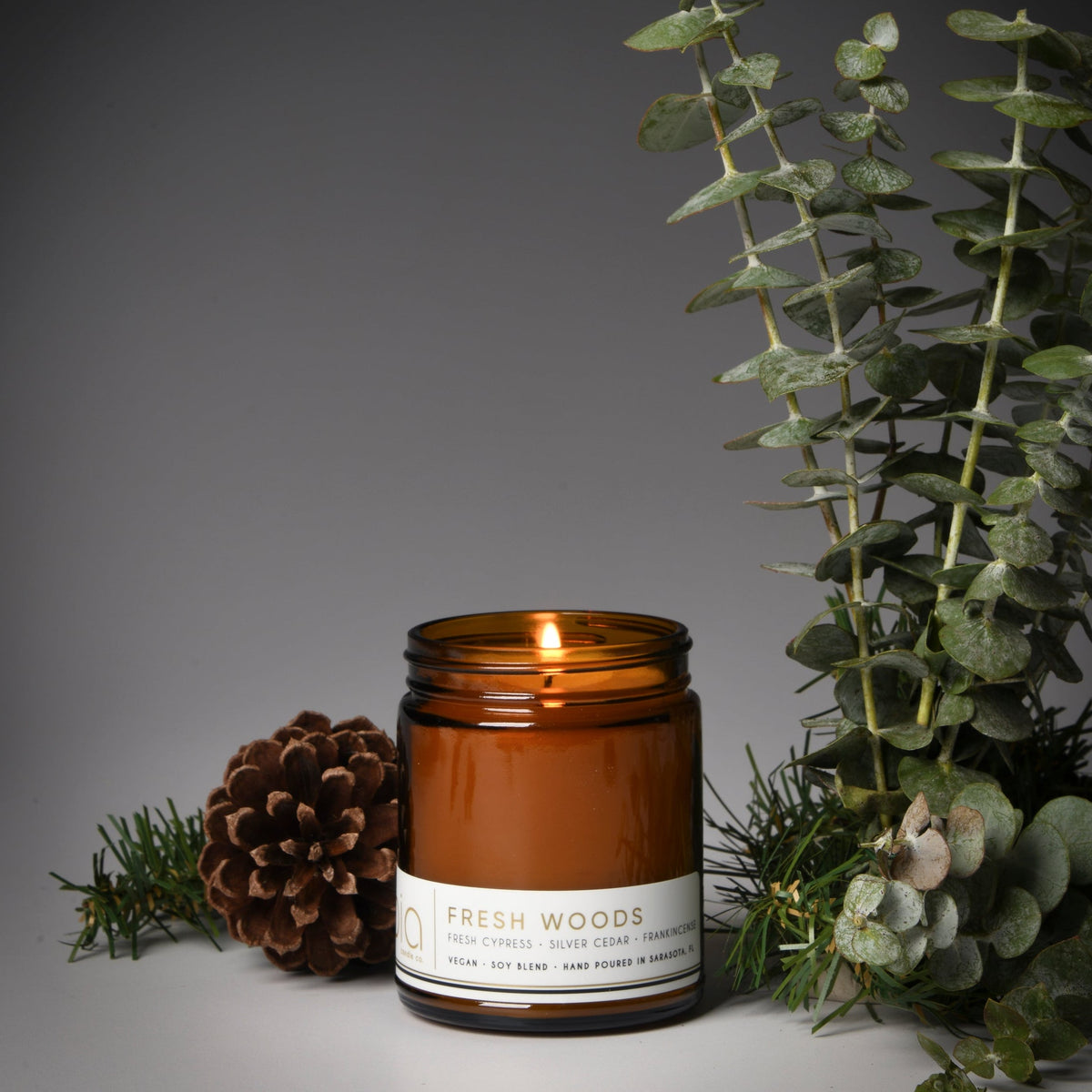 Lit soy blend candle jar with Fresh Woods label beside pinecones and green eucalyptus