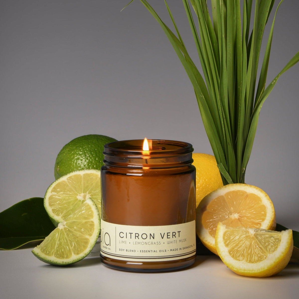 Citron Vert candle in amber jar with flame, surrounded by lemons and green lemongrass