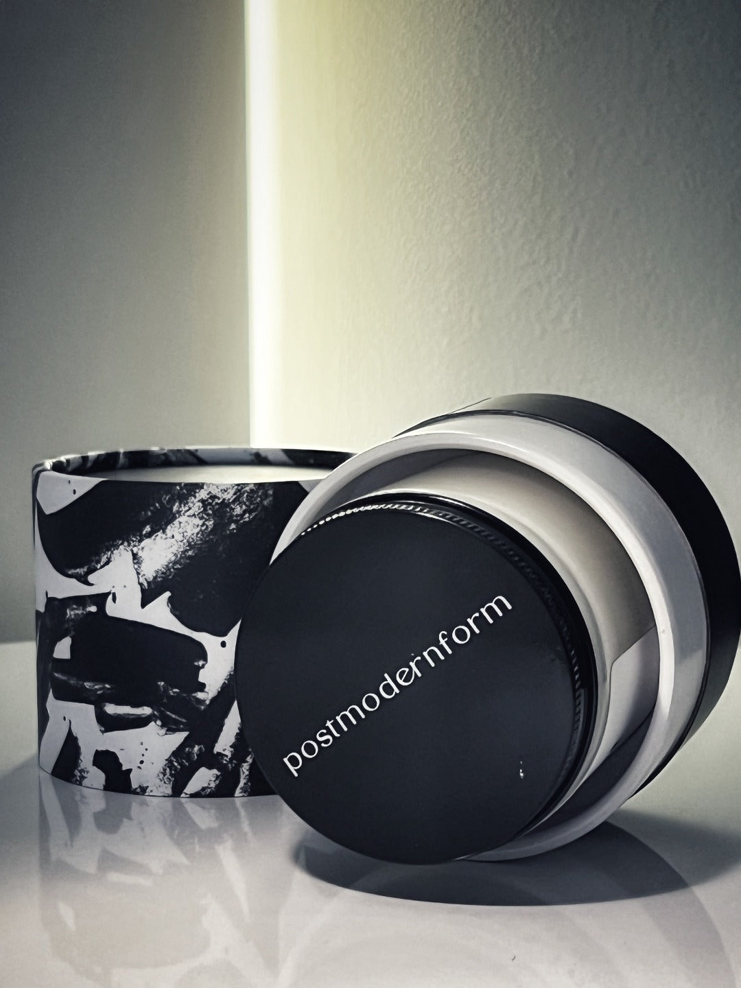 Candle jar with black lid and black-and-white printed sleeve, lid tilted beside jar