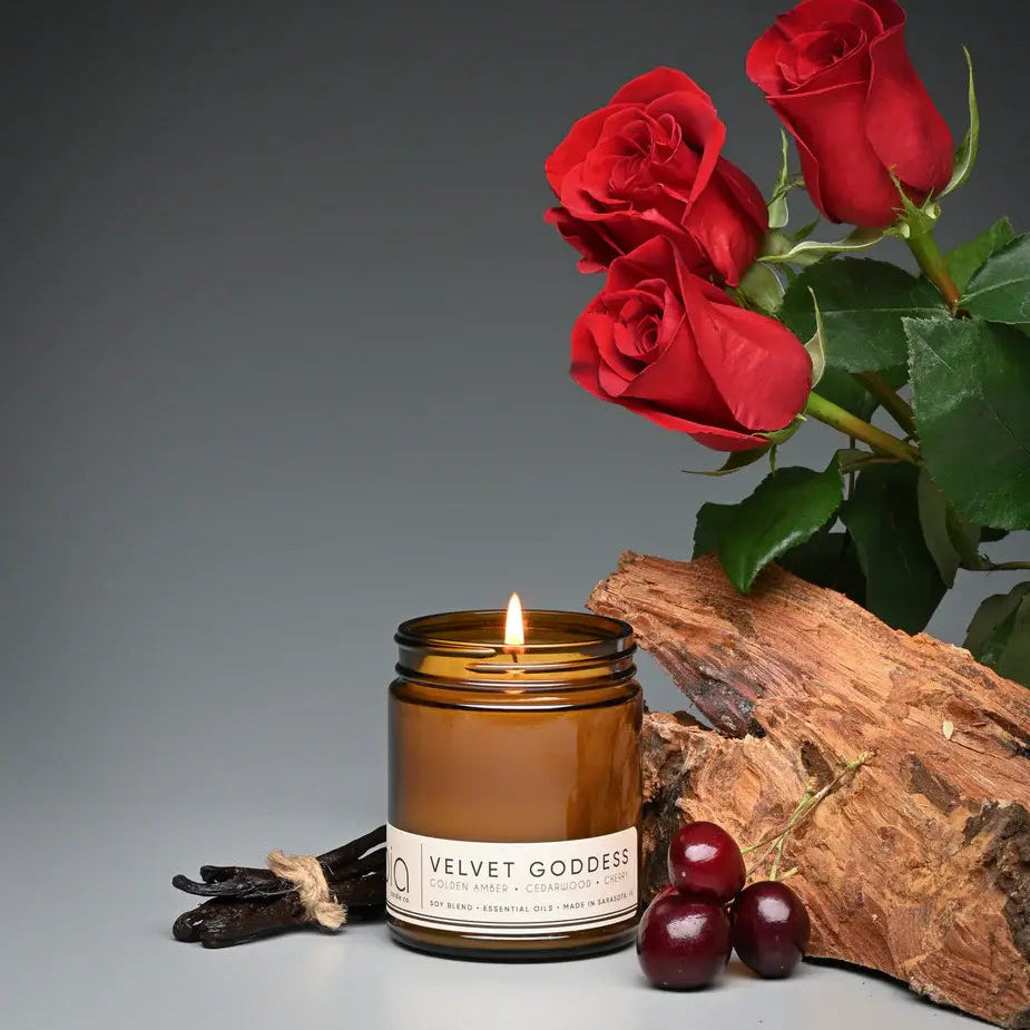 Amber glass soy candle with lit wick beside red roses and wooden log