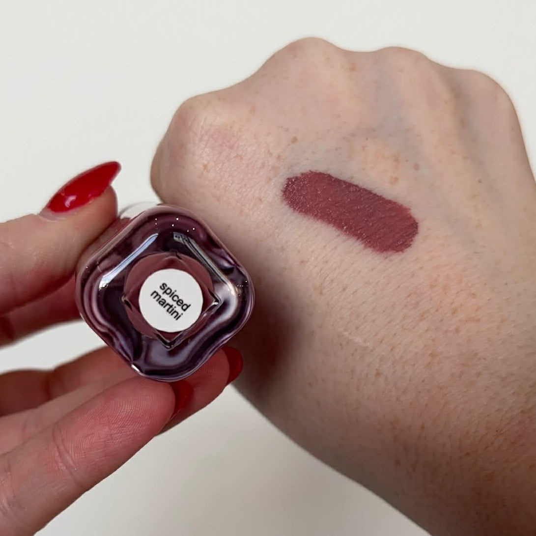 Hand holding a liquid blush bottle next to a dark mauve swatch on the arm.