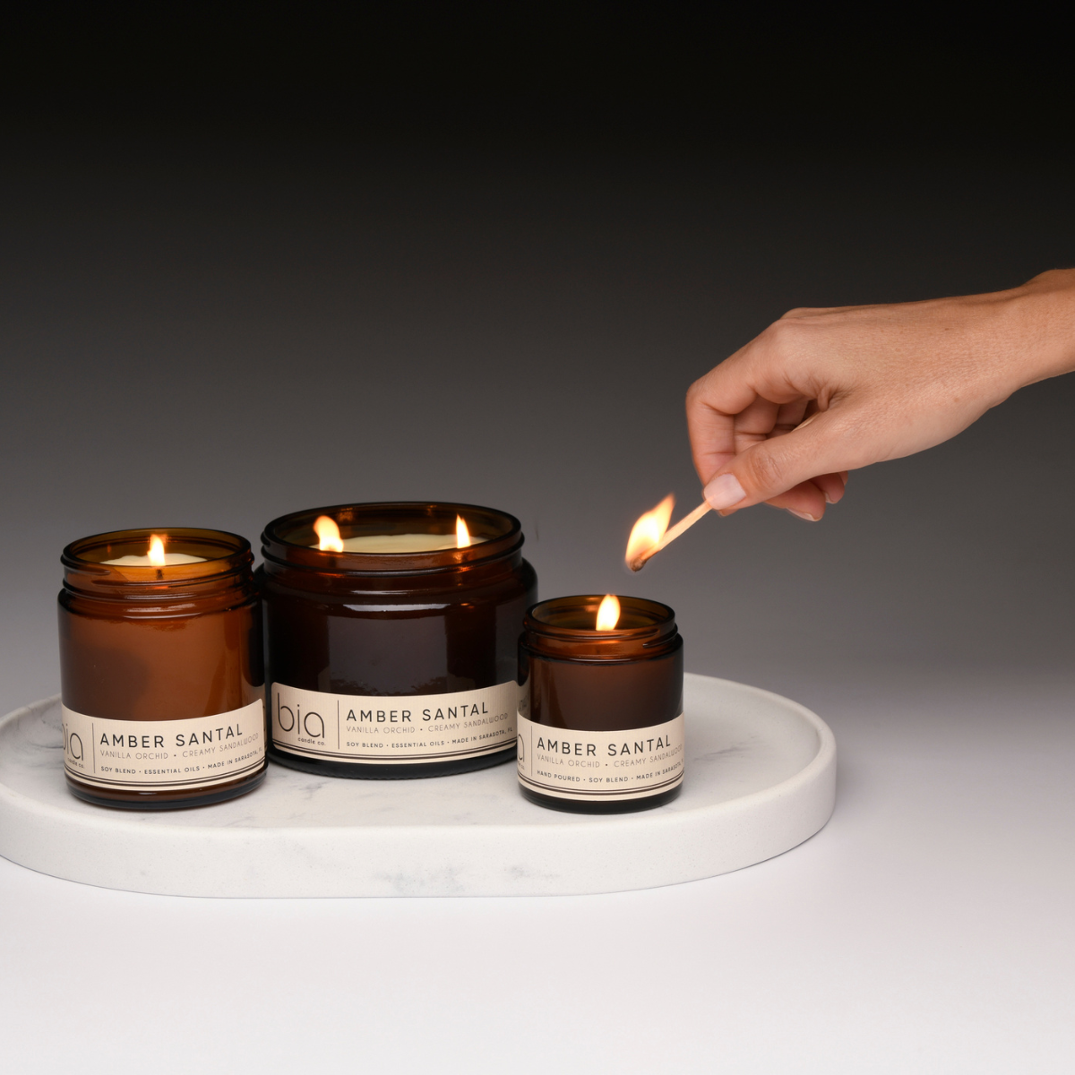 Set of amber glass soy candle jars on a tray; flames lit, hand holding a match.