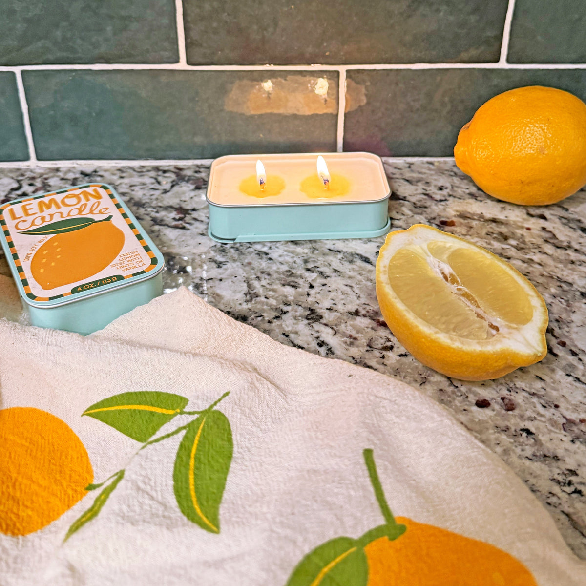Lit candle tin with two flames, with lemon fruit and towel on stone counter