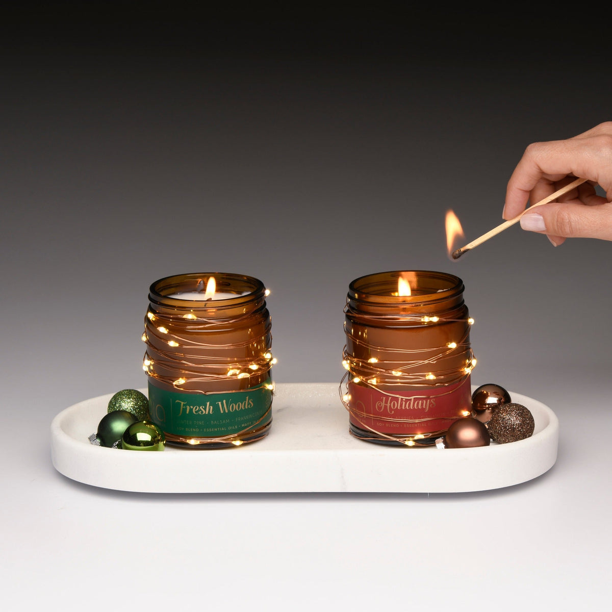 Two amber glass soy candles on a white tray, Fresh Woods (green) and Holidays (red), one lit