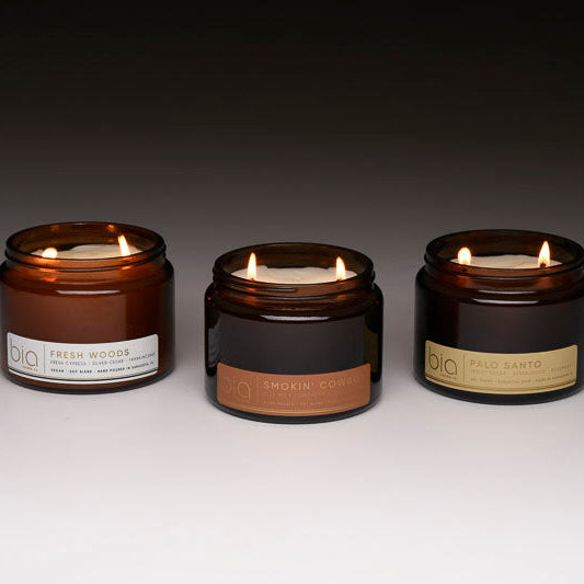 Three scented candles with flames in amber and dark glass jars on a flat surface.