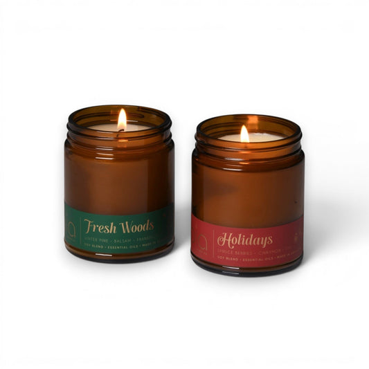 2 amber glass soy candles, one with green Fresh Woods label and one with red Holidays label