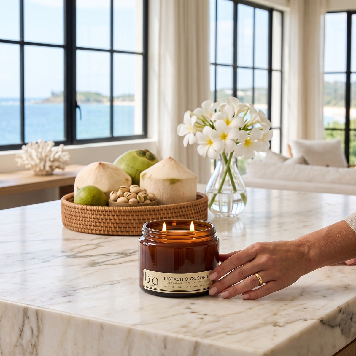 Amber candle jar with lit wick on marble countertop, hand holding jar beside flowers.