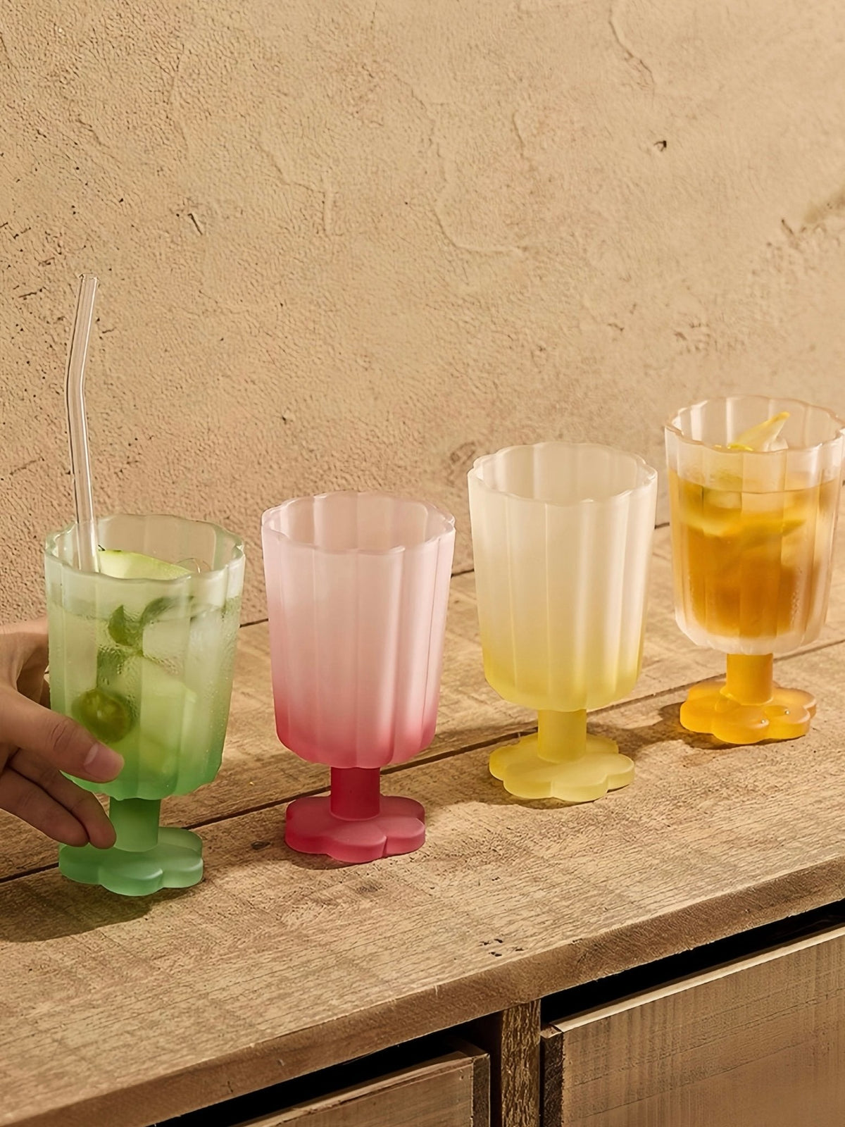 Hand holding a green hand-blown petal goblet, with pastel goblets on a wooden table.