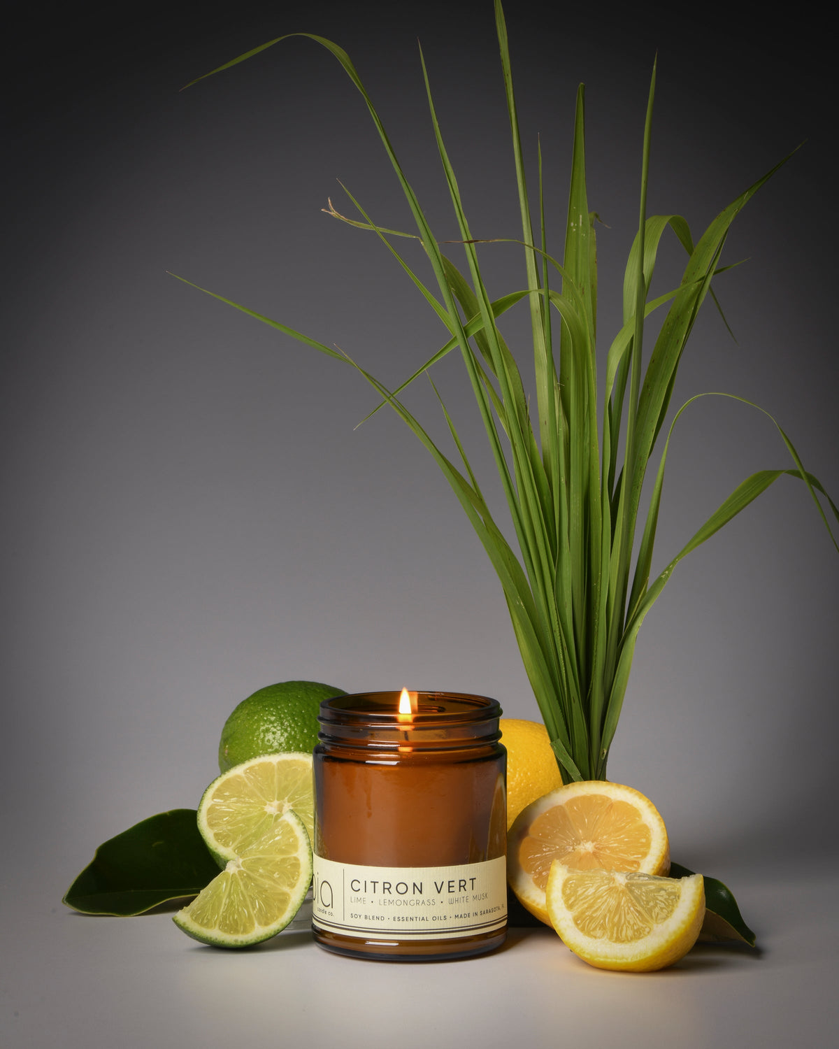 Citron Vert candle in amber jar with flame, with lemons and green lemongrass beside