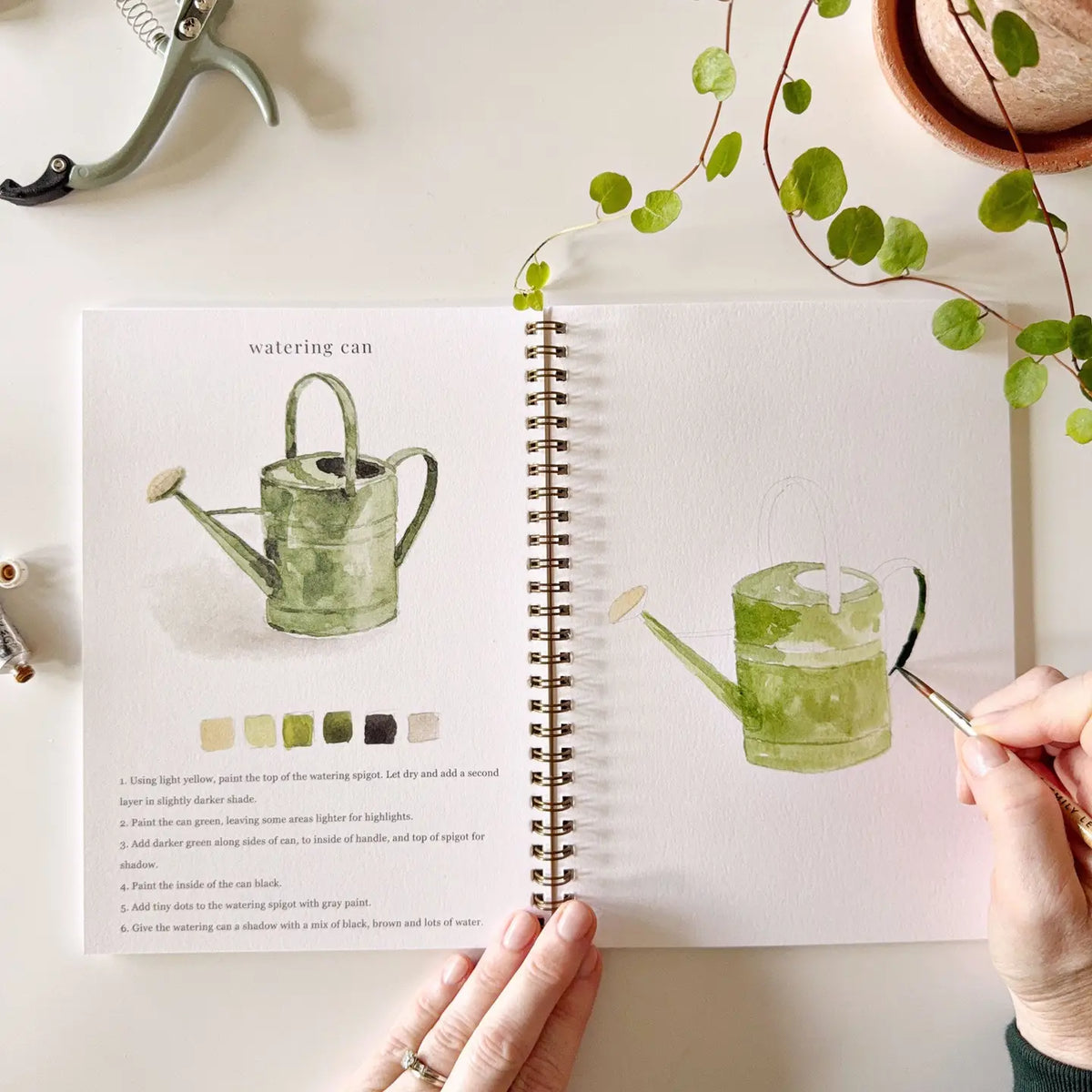 Garden watercolor workbook watering can painting with brush on right page, swatches on left