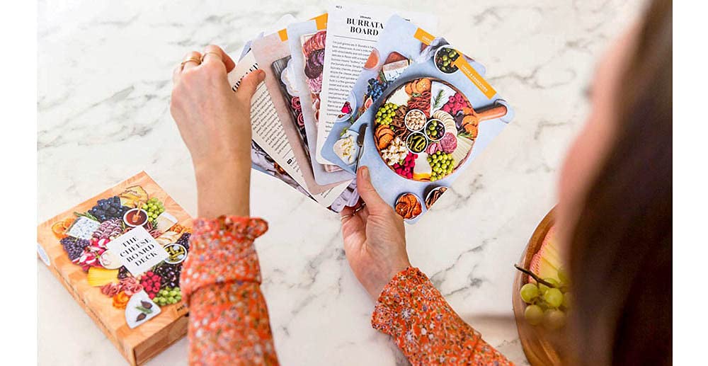Cheese board deck cards held in hands, showing colorful illustrated cheese spread themes