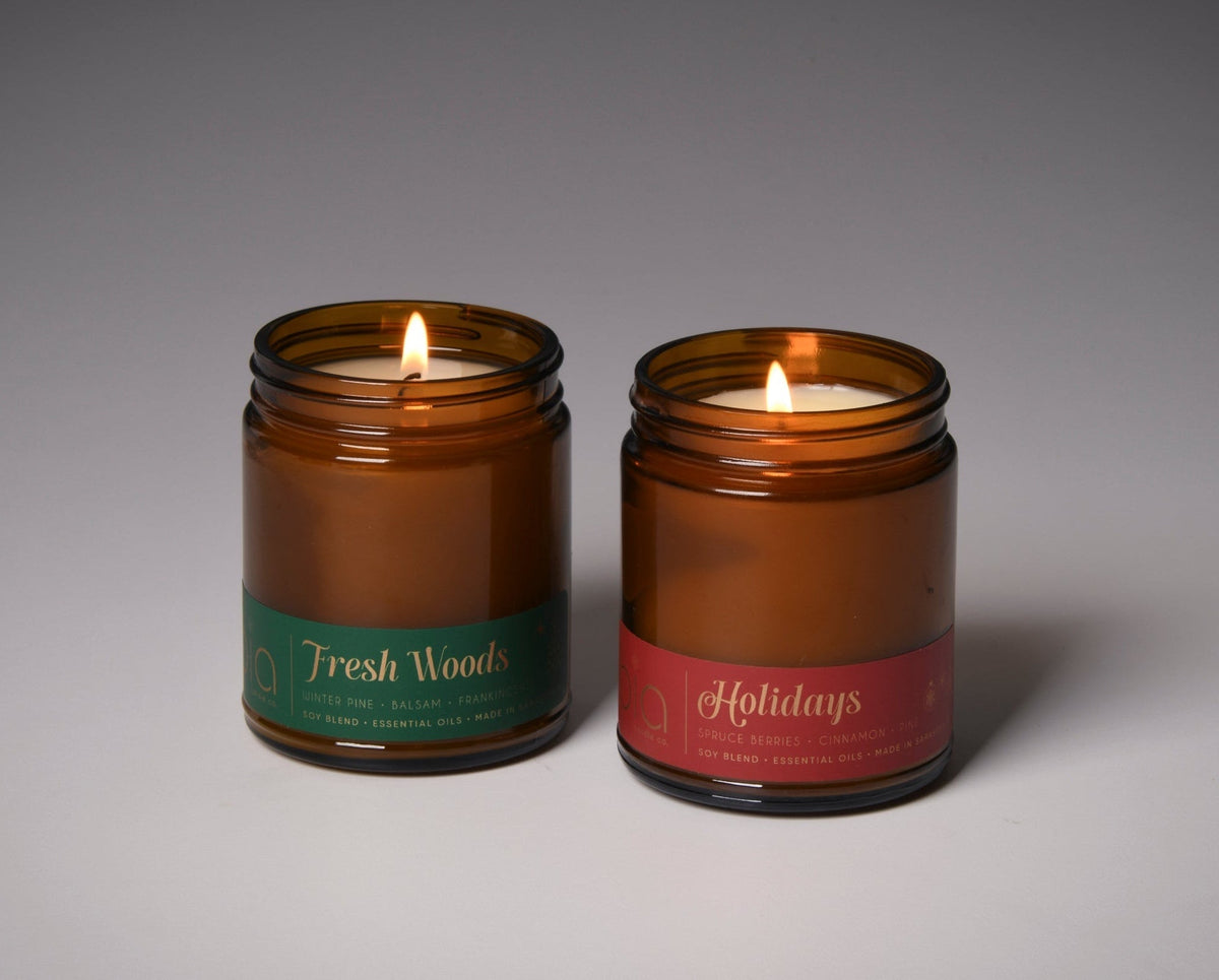 Set of two amber glass soy candles with green Fresh Woods and red Holidays labels, unlit