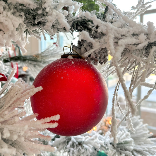 Red frosted glass ornament ball with hanging loop on a snow-dusted Christmas tree.