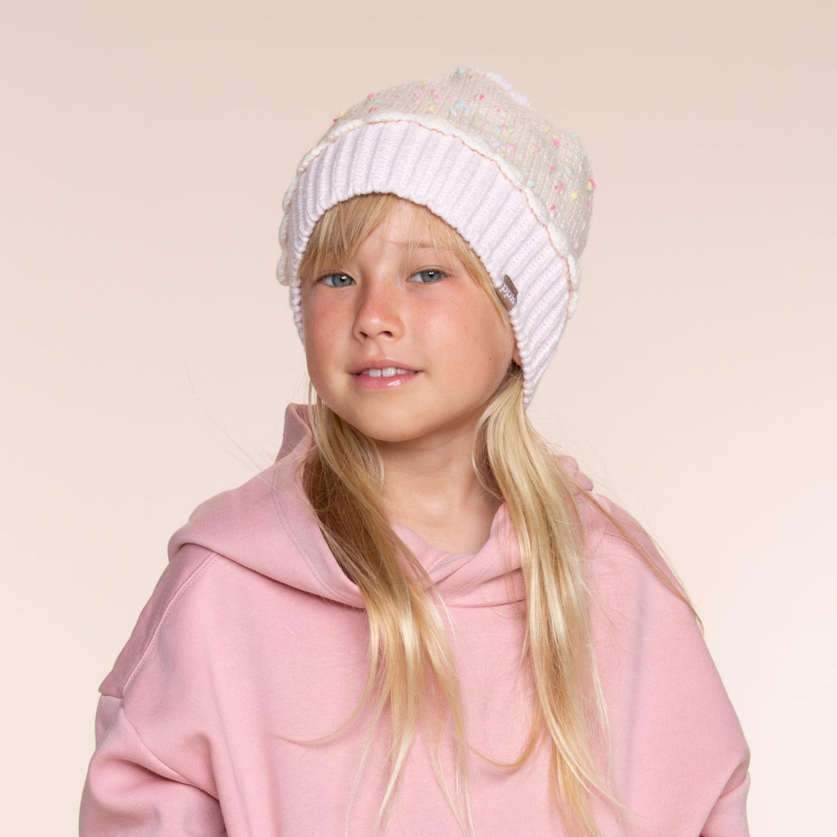 Cream sprinkle-knit children's beanie with pale pink ribbed cuff, worn with the cuff folded up