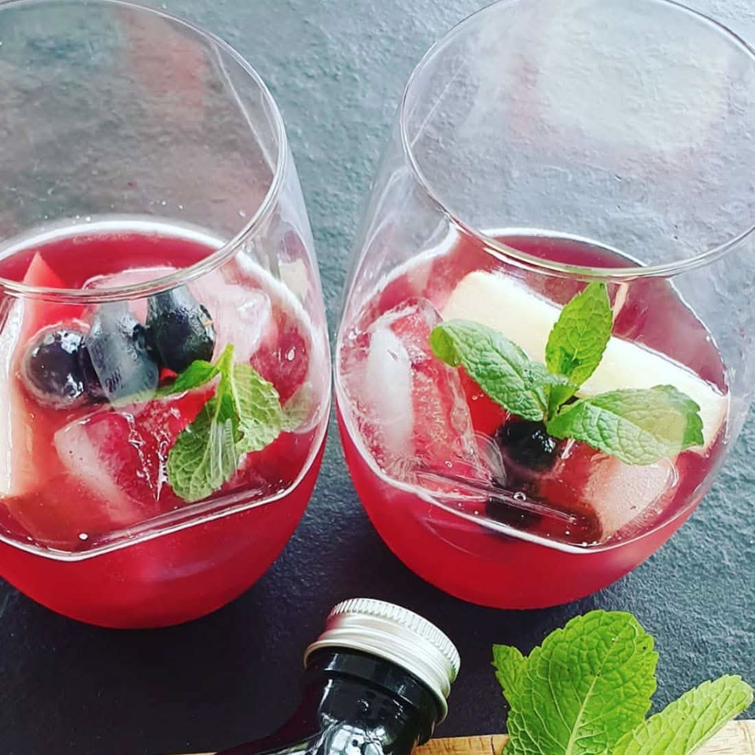 Two clear stemless wine glasses with red drink, ice, mint, and berries