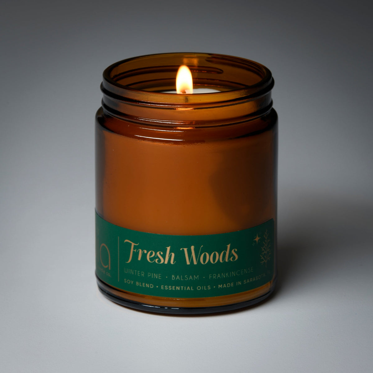 Amber glass candle jar with lit wick and Fresh Woods label in green text