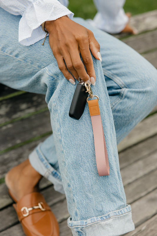 Pink wrist lanyard with tan patch, gold clasp, and black phone pouch in hand