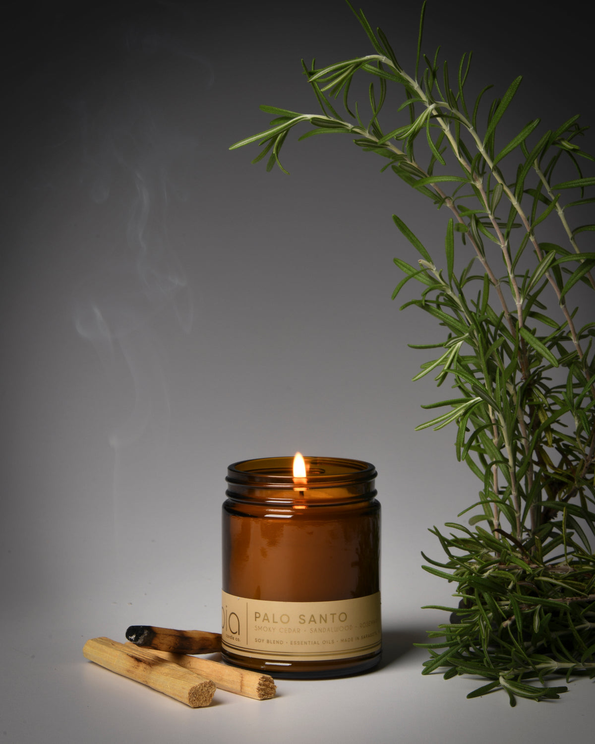 Amber glass soy candle with flame beside small wood sticks and green rosemary sprig