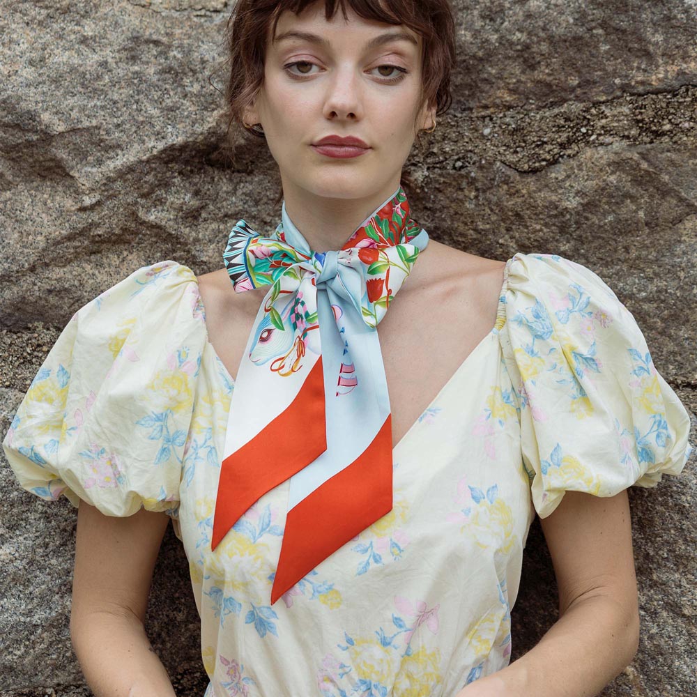 Silk twilly scarf worn at neck, light blue bird print with orange-red geometric tail