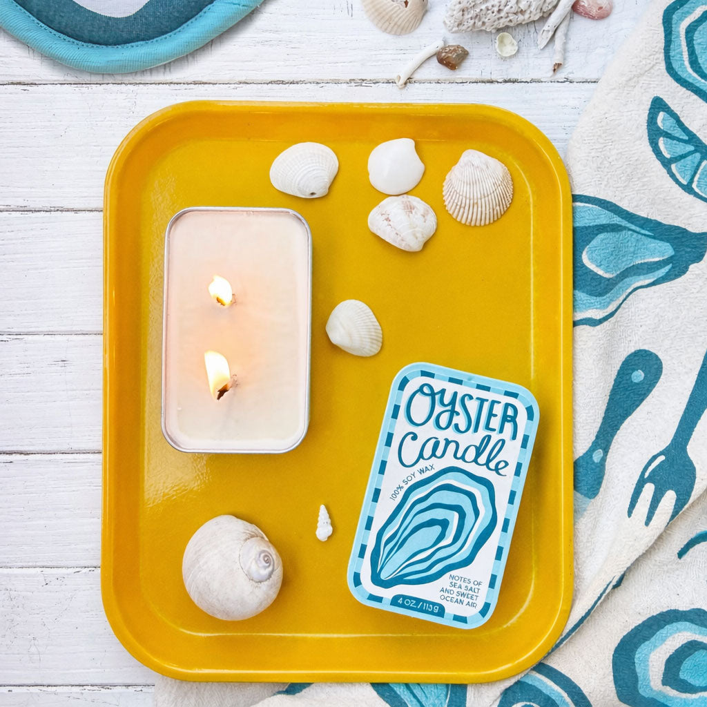 Oyster candle tin with flame in yellow tray, with shells and sea-themed towel