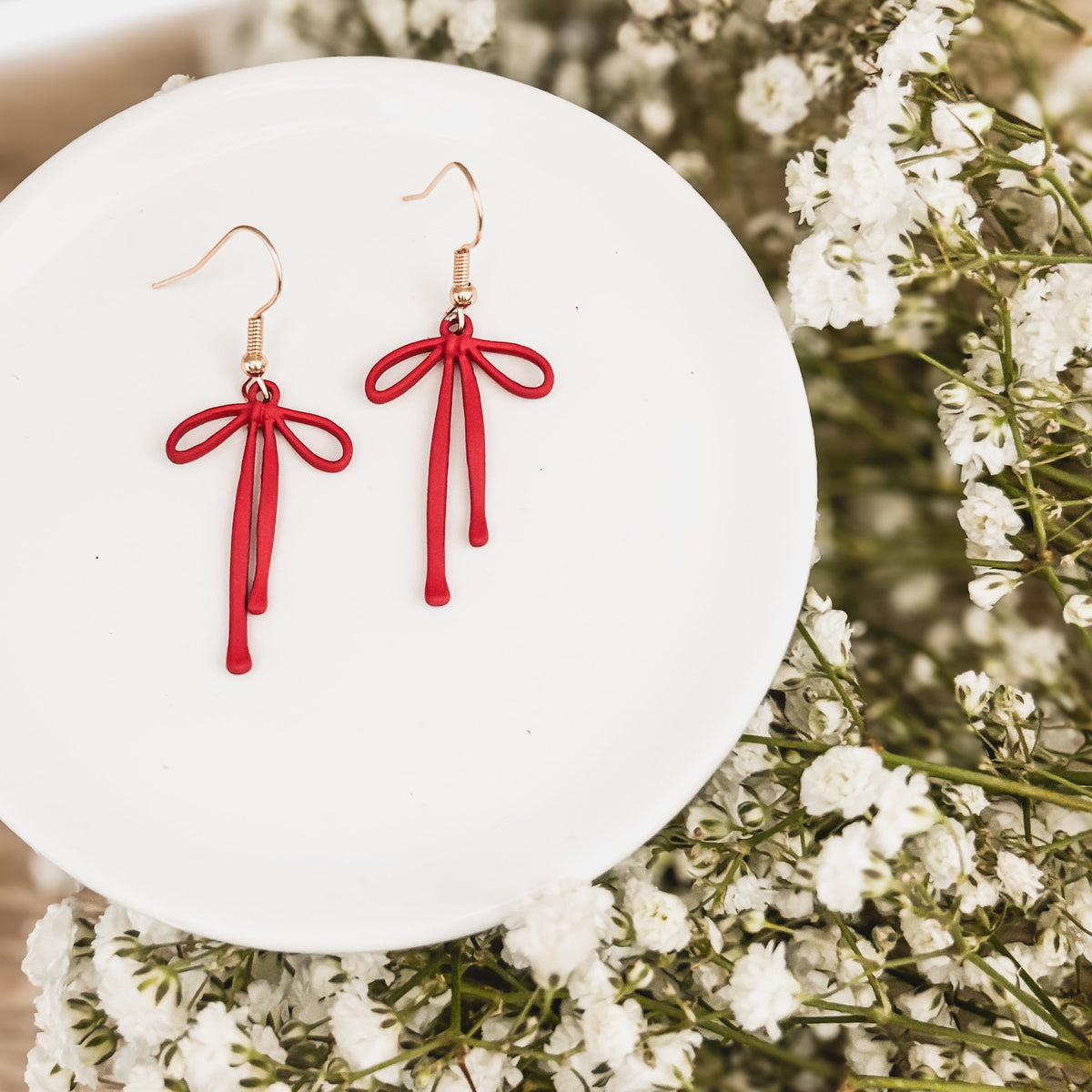 Pair of red bow drop earrings with dangling tassels and gold-tone hooks on a white plate