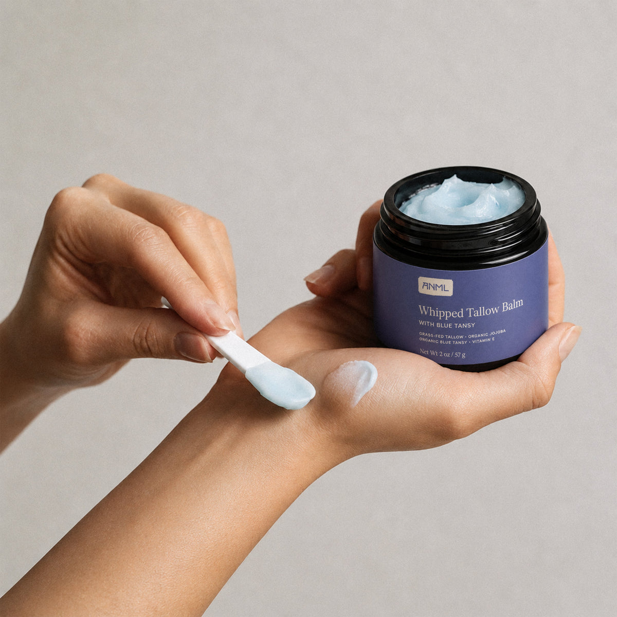 Hand holding a jar of whipped balm while applying a pale blue cream to skin