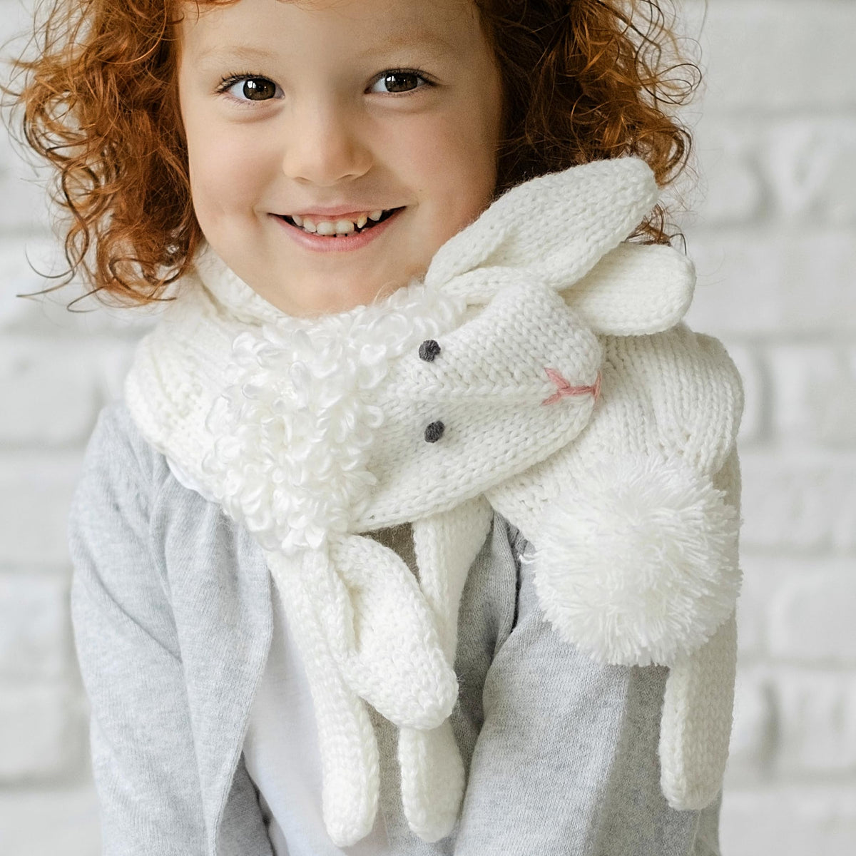 Child wearing a white knitted lamb scarf with a fluffy pom-pom and dangling legs