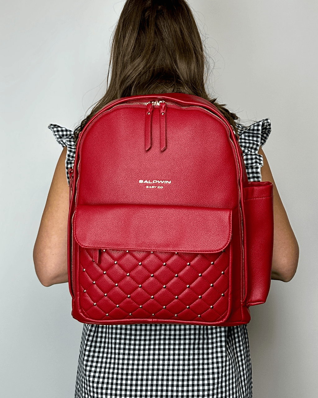 Crimson diaper backpack worn on the back, with a front flap and gold-studded quilted pocket.